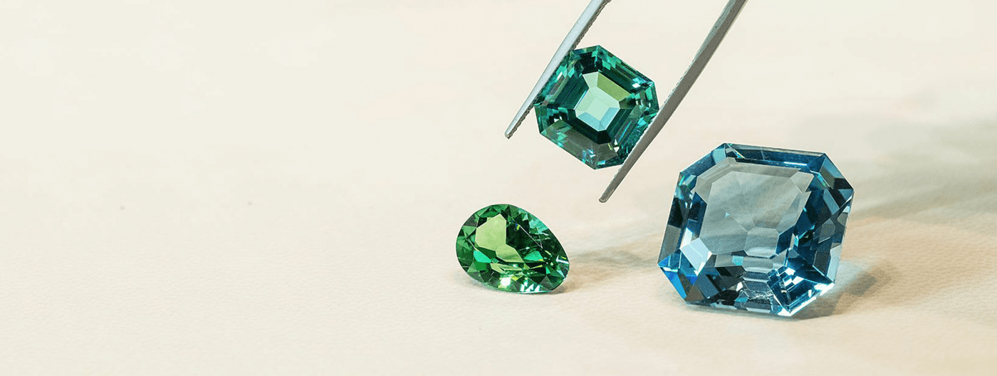 A green emerald &ndash;cut gemstone held by a pair of tweezers above a large blue octagon-cut gemstone and a pear-cut green stone.